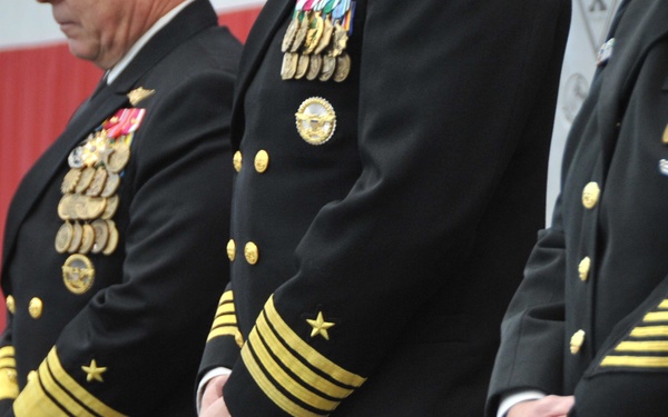 Three U.S. Navy officers in dress blue uniforms are standing at attention or in a formal posture, likely during a military ceremony. Their sleeves display gold rank stripes indicating senior officer grades, and their chests bear extensive medal and ribbon arrays. An American flag is partially visible in the background.
