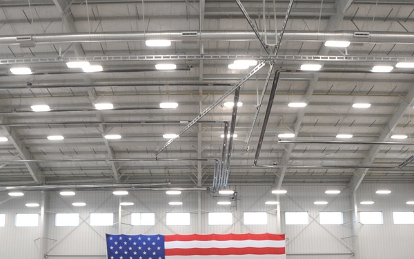 A formal military ceremony is taking place inside a large industrial hangar or facility. A large American flag is prominently displayed on the back wall as a backdrop, with a speaker at a podium addressing rows of seated military personnel. The audience is arranged in two sections facing the front stage area.
