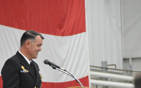 A senior U.S. Navy officer in dress blues is speaking at a wooden podium during a formal military ceremony. A large American flag serves as a backdrop, and an audience is partially visible in the foreground. The setting appears to be an indoor military facility or hangar.