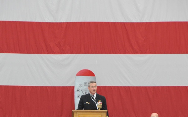 A formal U.S. Navy military ceremony is taking place in what appears to be a hangar or large indoor venue. A naval officer in dress uniform is speaking at a wooden podium bearing a shield/crest emblem, flanked by seated officers and officials in front of a large American flag backdrop. An audience of civilians and military personnel is seated in the foreground.