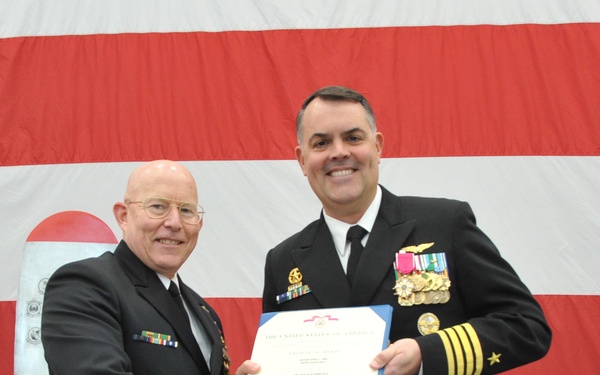 Two U.S. Navy officers in dress uniforms are shaking hands while jointly holding a certificate or award document. The ceremony appears to be a formal military recognition event, taking place in front of a large American flag backdrop. The officer on the right displays significantly more medals and insignia, suggesting a higher rank or the recipient of the award.