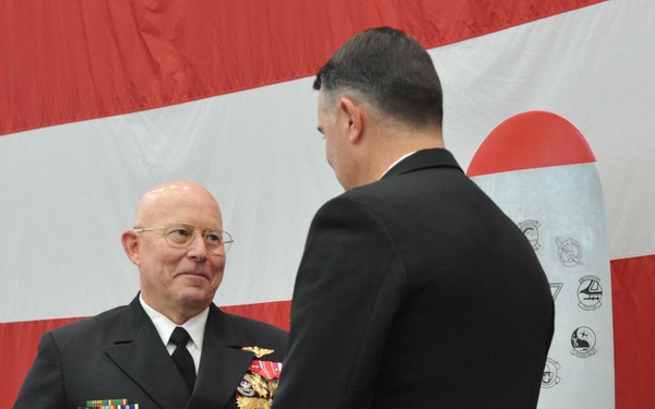 A formal U.S. Navy military ceremony is taking place in front of a large American flag backdrop. A senior naval officer wearing a dress uniform with multiple medals, ribbons, and gold sleeve stripes is interacting with another uniformed individual while holding a blue folder. The setting appears to be an awards or change-of-command ceremony.