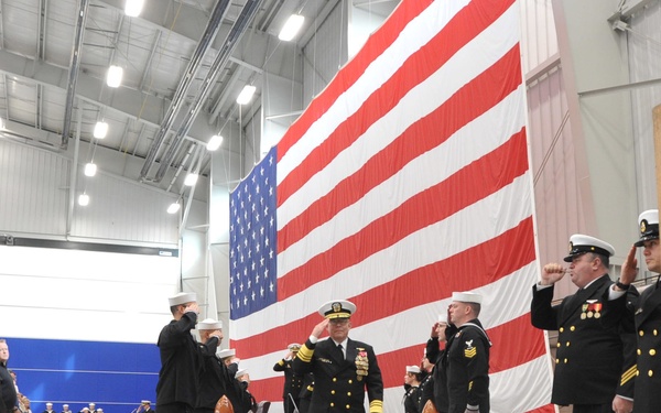 A formal U.S. Navy military ceremony is taking place inside a large indoor facility, likely a hangar or drill hall. A senior naval officer in dress uniform walks along a red carpet while personnel render salutes on both sides, with a large American flag displayed prominently in the background. A ship's bell on a wooden stand is visible in the foreground right, consistent with a change of command or commissioning ceremony.