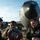 Four military personnel, appearing to be U.S. Air Force members based on their flight suits and leather jackets, are gathered on an airfield tarmac examining a worn object. They are standing in front of a large military aircraft, identifiable as a KC-135 tanker based on its distinctive nose profile and markings. The group appears engaged and in good spirits during what looks like a post-mission or pre-mission briefing moment.