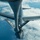 An aerial refueling operation is captured from the perspective of a tanker aircraft looking down at a B-52 Stratofortress bomber. The B-52 is positioned directly below the tanker's refueling boom, which is inserted into the bomber's receptacle. The image is taken at high altitude over an ocean or large body of water with scattered cloud cover visible below.