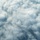 An aerial photograph captures a large multi-engine military aircraft flying above or alongside a dense cloud layer. The aircraft is positioned in the lower-left portion of the frame, with the vast majority of the image dominated by billowing white and grey cumulus clouds. The perspective suggests the photo was taken from another aircraft flying at a similar or slightly higher altitude.