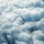 An aerial photograph showing a large multi-engine military bomber aircraft flying among dense cloud formations. The aircraft appears to be a B-52 Stratofortress, identifiable by its distinctive swept wings and multiple underwing engine pods. The image is taken from another aircraft at a similar altitude, providing a dramatic perspective of the bomber navigating through cumulus clouds.