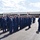 A formal United States Coast Guard muster or ceremony is taking place on a tarmac in front of a Coast Guard hangar facility. Personnel in dress blue uniforms and white combination caps are standing in formation while officers appear to be conducting an inspection or review. The tropical vegetation and architecture suggest this is likely a Coast Guard Air Station in Hawaii.