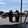 A military honor guard is carrying a flag-draped casket across an airport tarmac in what appears to be a dignified transfer ceremony. The casket is draped with an American flag, indicating the remains of a U.S. service member or dignitary. Personnel in dress uniforms are rendering salutes while a hearse is positioned nearby to receive the remains.