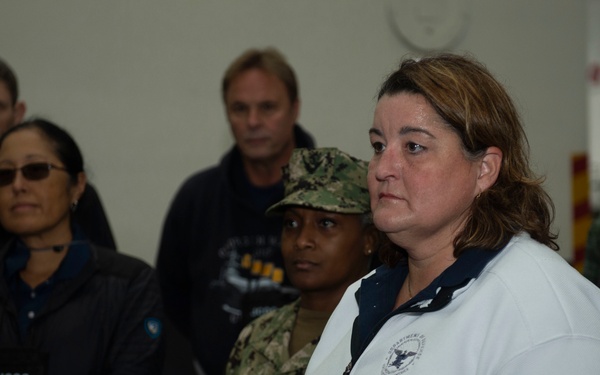 A group of individuals are gathered in what appears to be an official or military briefing setting. A woman in a white government-branded sweatshirt stands in the foreground holding printed materials with an American flag design. Several attendees are wearing 'JCOC' name badges, suggesting a Joint Civilian Orientation Conference or similar official program.