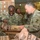 U.S. Army soldiers are handling MREs (Meals, Ready-to-Eat) in what appears to be a military warehouse or supply facility. Two soldiers in the foreground — one identified by nametape as 'BARTON' — are opening and inspecting MRE packages, while additional soldiers are visible in the background. The setting suggests a logistics or supply distribution operation.