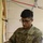A U.S. Army soldier with the last name 'Khan' is working with electrical wiring or cables in what appears to be a construction or installation setting. He is wearing Operational Camouflage Pattern (OCP) uniform with a unit patch on his right shoulder and 'U.S. ARMY' tape on his chest. The soldier is focused on handling wires or connectors near a wooden frame structure.