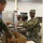 Military personnel are engaged in a field kitchen food service operation inside what appears to be a temporary or forward operating base dining facility. Soldiers in OCP (Operational Camouflage Pattern) uniforms are serving and receiving food, with a cardboard box labeled 'UGR-A' (Unitized Group Ration - A) visible in the foreground. The scene depicts a standard military meal service with food handlers wearing protective gloves.