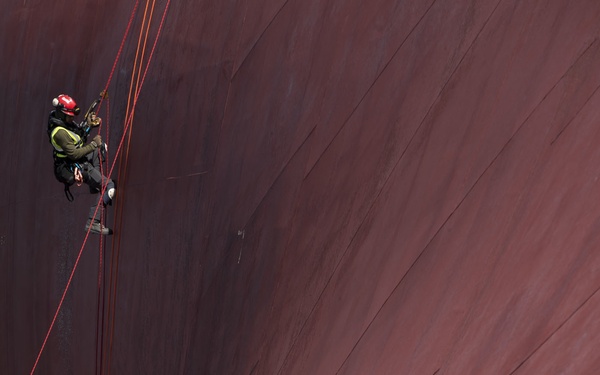 Two workers in safety harnesses and red helmets are suspended by ropes against the massive red-painted hull of a large ship. The hull dominates the frame, curving dramatically and showing signs of wear including scratches, rust patches, and surface damage. The scale contrast between the workers and the vessel emphasizes the enormous size of the ship.