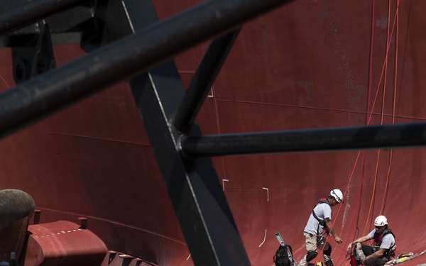 Two workers in safety harnesses and white helmets are performing maintenance or inspection work on the red hull of a large ship, likely in a dry dock or harbor setting. The image is captured through a foreground of black steel structural framework, which partially obscures the view. The workers appear to be suspended by ropes against the curved red hull near the stern of the vessel.