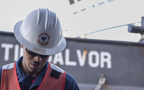 A U.S. Coast Guard National Strike Force member stands in front of a vessel named 'Atlantic Salvor' while operating a handheld device. The individual is wearing full personal protective equipment including a white hard hat bearing the National Strike Force Coast Guard Operations Team insignia and a red safety vest. The scene appears to be a maritime operational or salvage environment.