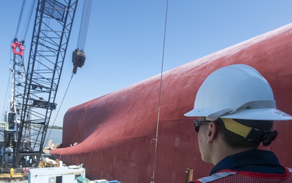 A U.S. Coast Guard personnel wearing a red safety vest and white hard hat observes a salvage or marine response operation from an elevated vantage point. A large red-hulled vessel, appearing to be capsized or listing severely, dominates the background while a crane barge with equipment and supplies is positioned alongside it. The scene suggests an active maritime emergency response or salvage operation.