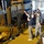 A group of military personnel from multiple nations are gathered on the deck of a naval vessel around large yellow industrial equipment, likely a launch and recovery system or winch mechanism. One individual in civilian-tactical attire is pointing upward, appearing to brief or instruct the group. Personnel are wearing a mix of US Navy, Australian Defence Force camouflage, and dark naval uniforms.