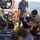 Military personnel from multiple nations are gathered on the deck of a naval vessel examining what appears to be an underwater mine or submersible device. A person in civilian-style dark clothing is pointing and explaining something about the equipment to the group. The setting appears to be a multinational naval exercise or training operation at sea.