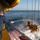 A worker aboard an offshore vessel is using a high-pressure water hose to wash down a piece of heavy yellow subsea equipment being recovered from the ocean. The equipment, suspended by multiple wire cables from a crane, appears to be a seafloor sampling or drilling tool being retrieved over the stern or side of the ship. The scene takes place in open ocean under clear skies.