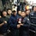 A group of military personnel, appearing to be a mix of Korean naval/military members and at least one Western individual, are gathered in a confined space that resembles the interior of a naval vessel or military facility. Two individuals in the center are shaking hands while jointly holding a commemorative shield/plaque, suggesting an official exchange or award ceremony. The group appears celebratory, with some members smiling and giving thumbs-up gestures.