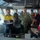 A group of military and civilian personnel are gathered on the bridge of a naval or maritime vessel. The group includes individuals in US Navy digital camouflage uniforms, Australian Defence Force camouflage uniforms, civilian high-visibility workwear, and emergency/safety red jackets. They appear to be conducting a briefing, inspection, or operational review on the ship's bridge.
