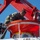 A person in military camouflage uniform is inspecting or working on a covered lifeboat or rescue craft marked 'LR5', suspended by a red davit crane system. A second individual wearing a blue hard hat and dark clothing observes nearby. The scene appears to take place aboard a vessel under clear blue skies.