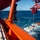 A maritime recovery or deployment operation is underway, with a white submersible or underwater vehicle labeled 'LRS FREMANTLE' being lifted or lowered into open ocean waters via a red crane/A-frame structure on a vessel. A person in military camouflage uniform and high-visibility vest with a white hard hat observes the operation from the ship's deck. A small inflatable rubber boat with personnel is positioned nearby in the water, likely providing safety or recovery support.