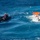 The image shows a maritime rescue or training operation in open blue ocean waters. A black rigid inflatable boat (RIB) with two crew members is positioned near a white and orange vessel that appears to be capsized or in distress, with personnel in the water alongside it. The scene suggests an active search-and-rescue or man-overboard drill exercise.