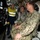 Three individuals are inside what appears to be a hyperbaric chamber or submarine pressure vessel facility. Two are wearing U.S. Navy camouflage uniforms (NWU Type III), while a third person in a high-visibility safety vest appears to be a civilian technician or guide. The group appears to be on an inspection or familiarization tour of the pressurized chamber equipment.