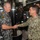 Two senior military officers — one from the Royal Australian Navy and one from the U.S. Navy — are shaking hands inside what appears to be a submarine interior. The Australian officer wears a patch identifying the vessel as HMAS Sheean (SSG 77). Several other naval personnel in Australian camouflage uniforms are visible in the background.