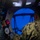 A military personnel member in digital camouflage uniform is observed at a control station, likely inside a naval vessel or diving/hyperbaric facility. The operator faces a circular porthole or pressure chamber hatch with a blue-lit interior visible beyond it. The control panel to the left features analog gauges, digital readouts, indicator lights, and various switches.