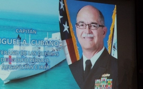 A large projection screen displays a military tribute slide featuring Captain Miguel A. Cubano, identified as a veteran of the Persian Gulf War, Afghanistan, and Iraq conflicts. The slide is in Spanish and shows a formal naval portrait alongside an image of a military vessel. The setting appears to be a formal ceremony or presentation event.