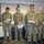 A group of U.S. Navy personnel in NWU Type III (digital camouflage) uniforms are gathered in an indoor room, most holding papers and appearing to read from them. An American flag is visible on the right side of the frame. The setting appears to be an official military ceremony or administrative event, possibly a re-enlistment, promotion, or oath-taking ceremony.