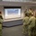 A group of U.S. Navy personnel in NWU Type III (woodland digital camouflage) uniforms are gathered in a briefing room attending a presentation. A projected screen displays an image of ocean waves, suggesting a maritime or beach-related briefing. Several personnel are holding papers and appear to be actively participating in the session.