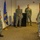 A group of U.S. Navy personnel in NWU Type III (woodland camouflage) uniforms are gathered in what appears to be a briefing or ceremony room. One sailor on the left appears to be addressing the group, while others stand attentively in a semi-circle formation. A blue and gold military flag and a podium with a naval command seal are visible in the background.