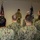 A group of U.S. Navy personnel in NWU Type I (digital camouflage) uniforms are gathered in an indoor space, facing a single service member who appears to be speaking or reading from a document. Two flags are visible in the background — an American flag on the left and what appears to be a Navy command flag on the right. The setting suggests a formal military ceremony, briefing, or muster.