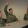 Three U.S. Navy personnel in NWU Type I (digital camouflage) uniforms are seated at a table in what appears to be a briefing or classroom setting. The center individual, identified by name tape as 'GEORGE,' has their right hand raised, likely asking a question or responding to a prompt. An American flag is visible in the background to the right.