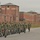 A large formation of military personnel in camouflage uniforms marches in organized columns along a paved road at what appears to be a military installation or naval base. The group is led by a flag bearer carrying a guidon, with a traffic control personnel in an orange vest observing from the side. Red brick buildings and fencing characteristic of a military base are visible in the background.