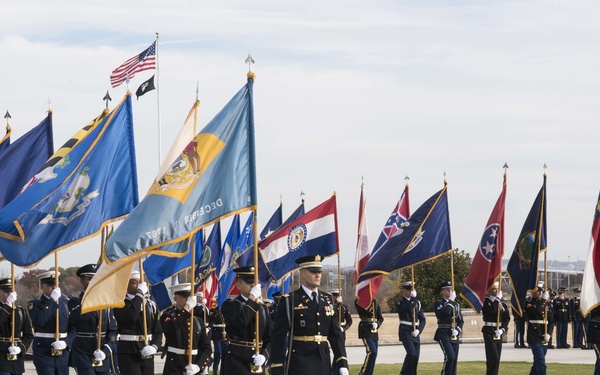 A formal military color guard ceremony is taking place on an open grassy field, with uniformed service members carrying a large array of U.S. state and military flags. The personnel are dressed in formal dress uniforms representing multiple branches of the U.S. military, marching in organized formation. An American flag and what appears to be a POW/MIA flag are visible on a flagpole in the background.