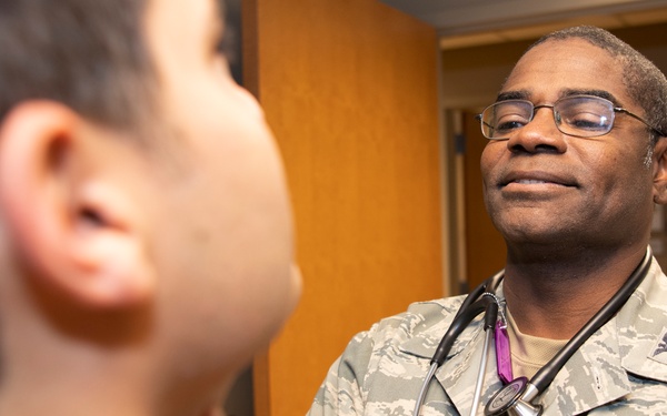 A U.S. Air Force medical officer named Britten, wearing ABU (Airman Battle Uniform) with a stethoscope, is conducting a medical examination of a patient. The officer appears to be palpating or examining the patient's neck/throat area in what appears to be a clinical examination room. The interaction suggests a routine medical consultation or physical examination.