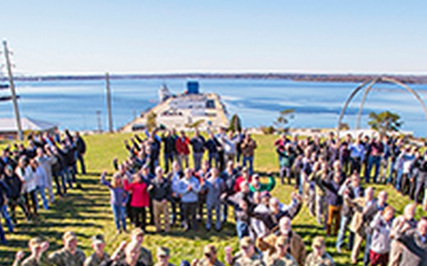 A large group of people, including several individuals in military camouflage uniforms, are arranged in a horseshoe or heart-shaped formation on a grassy lawn near a waterfront. The aerial or elevated perspective captures the organized group pose, with a pier, large vessel, and body of water visible in the background. Some participants are waving or raising their hands toward the camera.