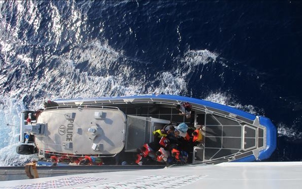 An aerial/overhead view from a larger vessel looking down at a blue and gray rigid inflatable boat (RIB) marked 'POLICE' alongside the ship. The police vessel appears to be carrying multiple people wearing life jackets or high-visibility gear. The foreground shows what appears to be a helipad deck with a painted 'H' symbol and a colorful flag display.