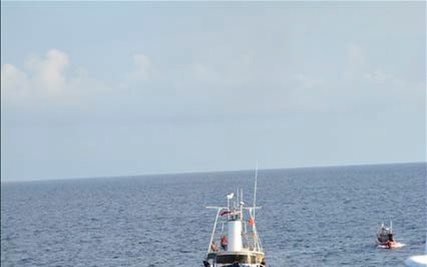 The image shows a maritime operation scene viewed from the flight deck of a large vessel, with an orange search-and-rescue helicopter (numbered '70') in the foreground. A blue and red fishing or patrol vessel is visible in the middle distance on open ocean, with a smaller red boat visible further in the background. The scene suggests a Coast Guard or maritime law enforcement interdiction or rescue operation.
