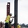 Three construction workers are engaged in steel beam erection on an elevated work platform. The foreground workers are positioning or aligning a vertical steel I-beam, while a third worker is visible in the background. All workers are wearing standard personal protective equipment including hard hats, high-visibility vests, and safety harnesses.