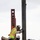 Three construction workers are actively engaged in steel erection work at an elevated construction site. The workers are positioning and securing a vertical steel column or beam, working in close coordination on what appears to be a bridge or large structural platform. All workers are equipped with standard personal protective equipment including hard hats, high-visibility vests, and safety harnesses.