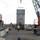 An active construction site showing workers overseeing the crane-assisted placement of a large steel panel or wall section. An orange crawler crane (marked 'CR 003') on the right is lifting the steel structure using a spreader bar. Multiple workers in high-visibility vests and hard hats are positioned around the lift zone, with additional heavy equipment visible in the background.