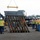 A construction site scene showing workers in high-visibility safety gear guiding a large steel structural assembly being lifted by a crane with a yellow spreader bar. Multiple workers wearing hard hats and reflective vests are managing tag lines attached to the steel frame structure. Heavy equipment including a wheel loader and other vehicles are visible in the background.
