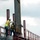 Two construction workers are positioned at elevation on a steel-frame structure, guiding or securing a large vertical steel beam or column. Both workers are equipped with personal protective equipment including hard hats, high-visibility vests, and fall protection harnesses. A cable or wire rope is visible, likely used for rigging or stabilizing the steel member being placed.