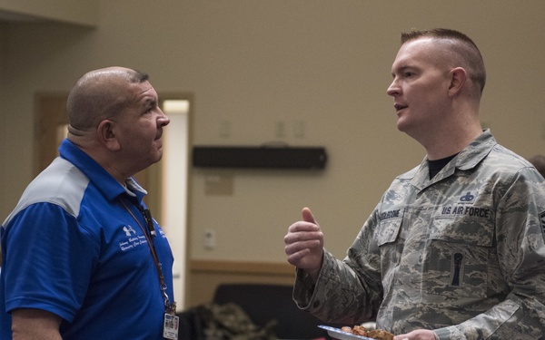 Two men are engaged in conversation in what appears to be a military dining or event facility. One man is wearing a US Air Force camouflage uniform with visible rank insignia (Senior NCO chevrons) and holding a plate of food, while the other is wearing a blue polo shirt with a lanyard/badge. The setting suggests a social or professional gathering event.